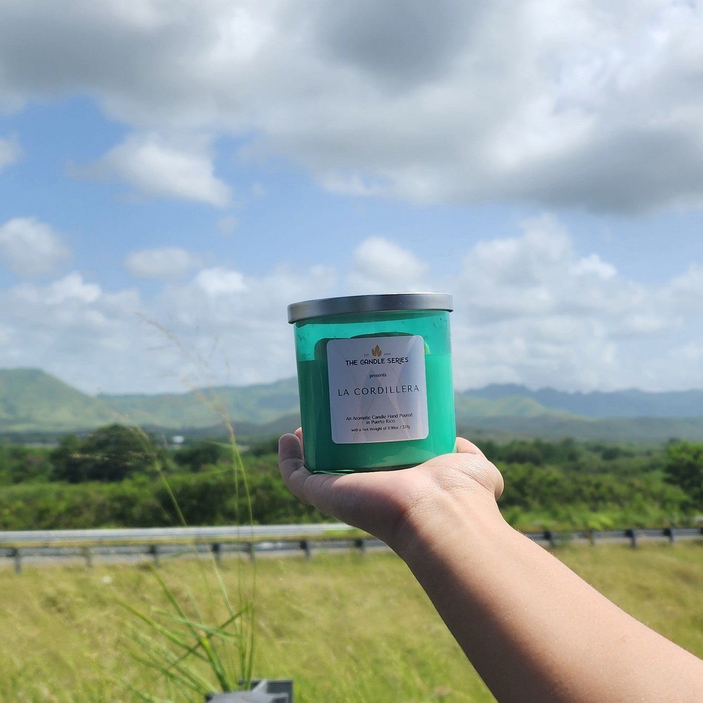 Hand holding a green candle against a scenic background with mountains and clouds.